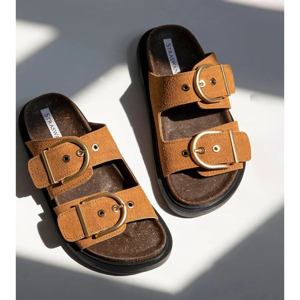 A pair of brown double-strap slide sandals with large gold-tone buckle details, displayed on a white surface with dramatic diagonal shadows.