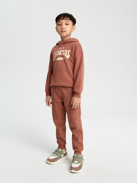 Child wearing a brown hoodie and pants set with 'ADVENTURE' text on a white background