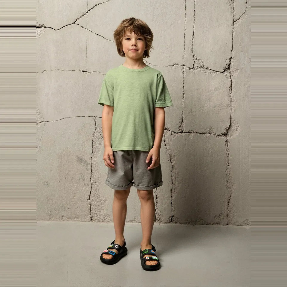 A young boy stands against a textured concrete wall wearing a light sage green short-sleeve t-shirt, gray shorts, and black slide sandals.