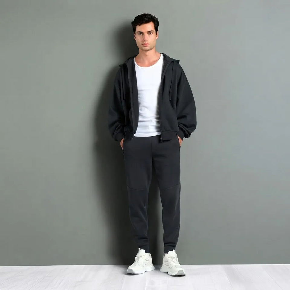 A man stands against a gray wall wearing a black zip-up hoodie over a white t-shirt, dark gray sweatpants, and white sneakers.