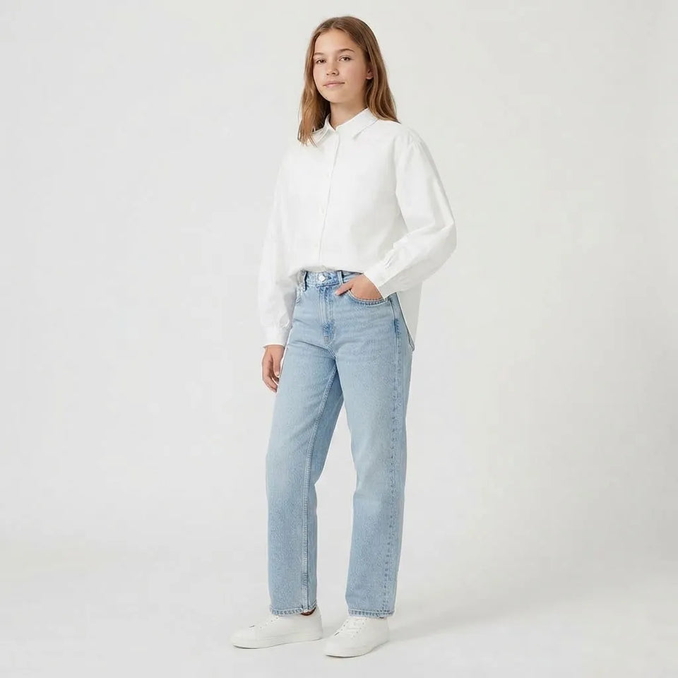 A young woman stands against a plain white studio background wearing a white long-sleeve button-up shirt, light-wash blue straight-leg jeans, and white sneakers.