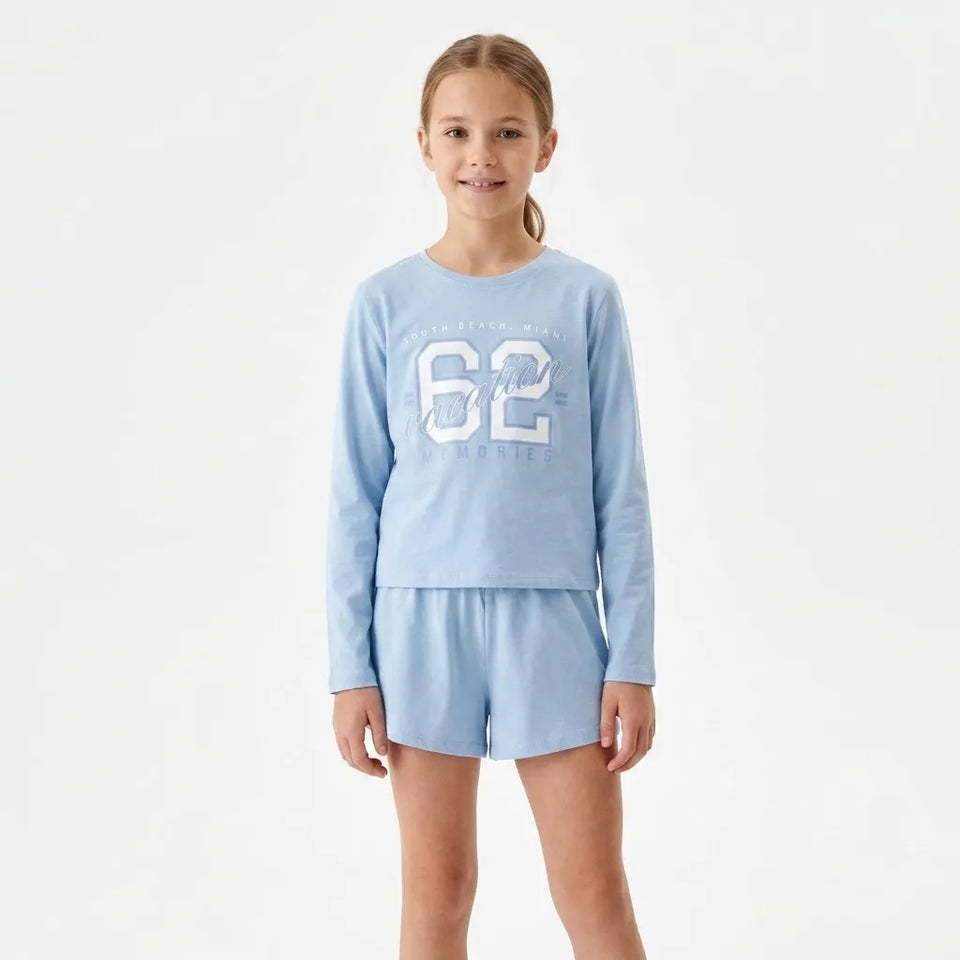 A young girl modeling a light blue two-piece pajama set consisting of a long-sleeve top with a large white '62' graphic and matching shorts, set against a plain white studio background.