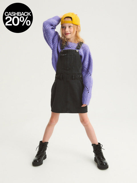 Person wearing a purple sweater, black denim overall dress, and yellow cap on a white background