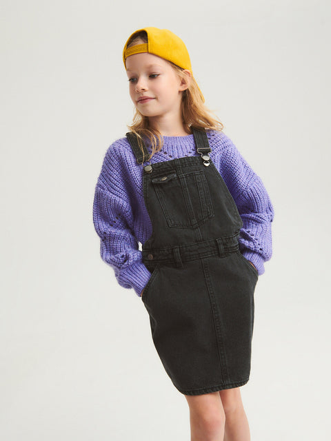 Child wearing a purple sweater and black denim overalls with a yellow cap on a light gray background