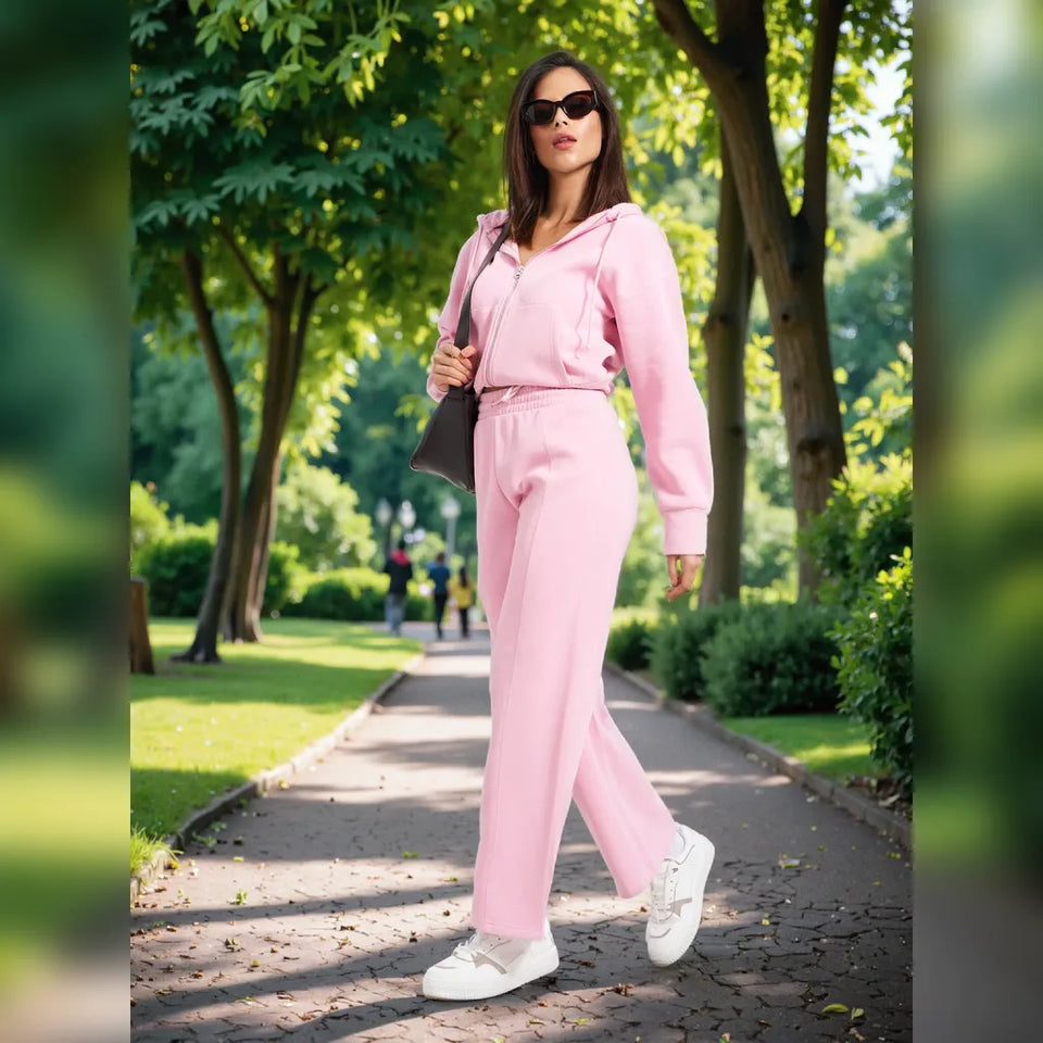 A woman wearing a light pink two-piece tracksuit with a zip-up hoodie and matching wide-leg pants, walking on a paved path in a sunny park. She is wearing white sneakers and carrying a dark gray shoulder bag.