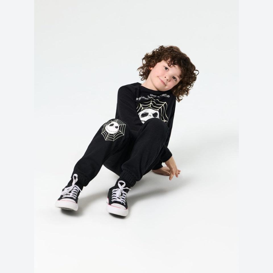 A child with curly brown hair sits on a light gray studio floor wearing a black long-sleeve pajama set with spider-themed graphics, including a spider face on the chest and spiderweb motifs on the sleeves, paired with black and white high-top sneakers.