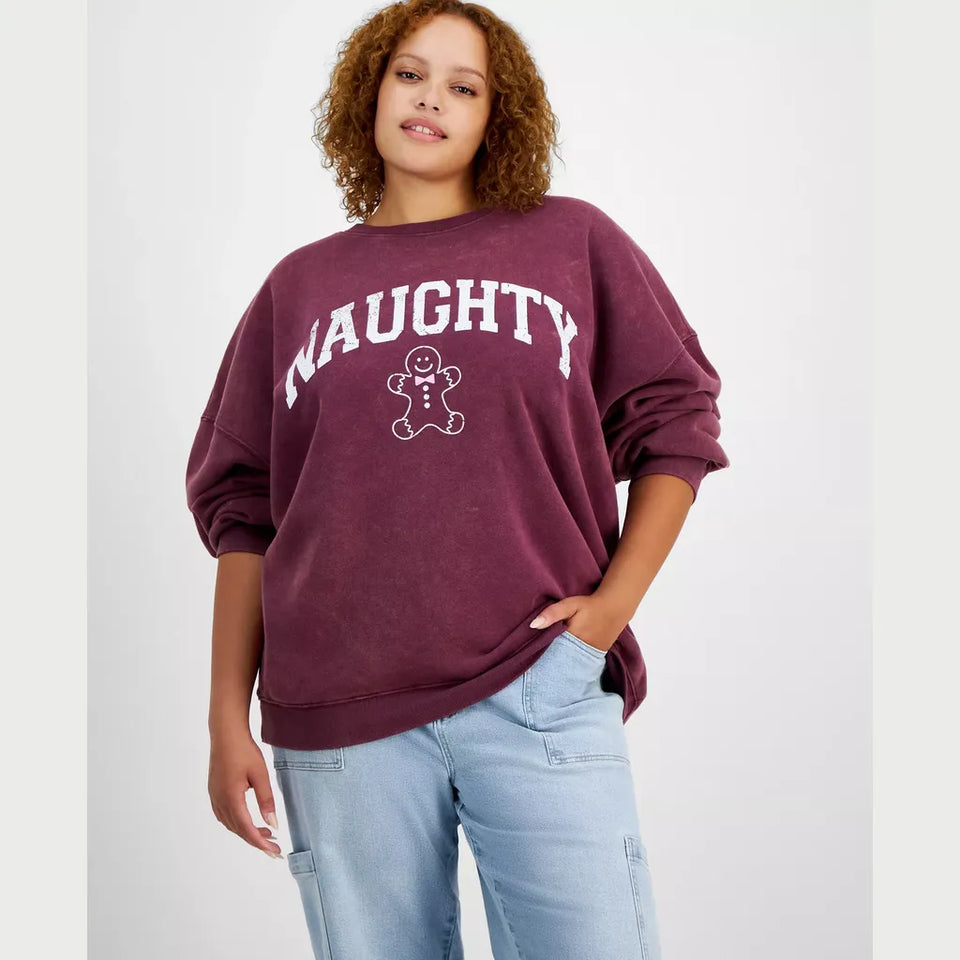 A woman wearing a maroon crewneck sweatshirt with 'NAUGHTY' and a gingerbread man graphic printed in white on the front, paired with light blue jeans, against a light gray studio background.