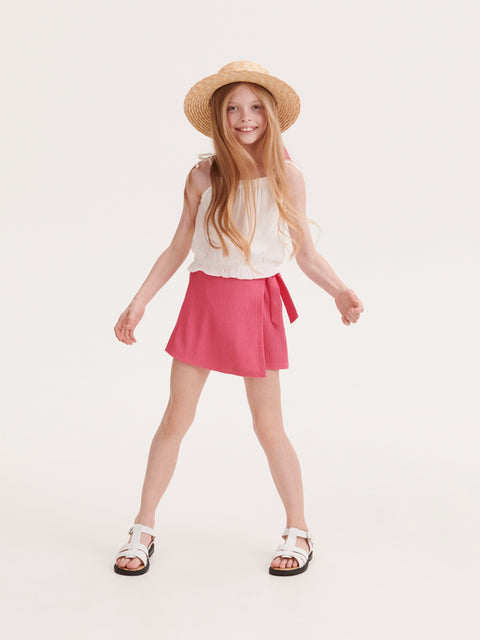 Young girl wearing a white sleeveless top and pink skirt with a straw hat on a white background