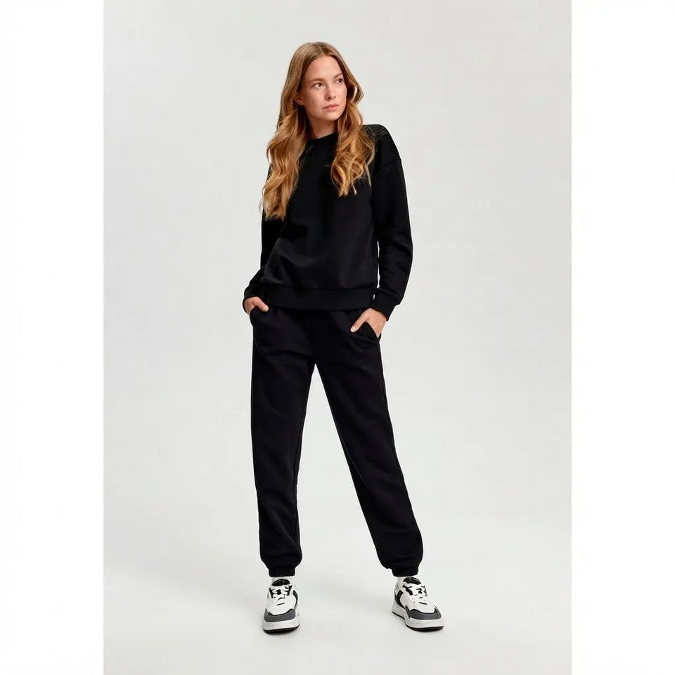 A woman modeling a black crewneck sweatshirt and matching black sweatpants with white and black sneakers, standing against a light gray studio background.
