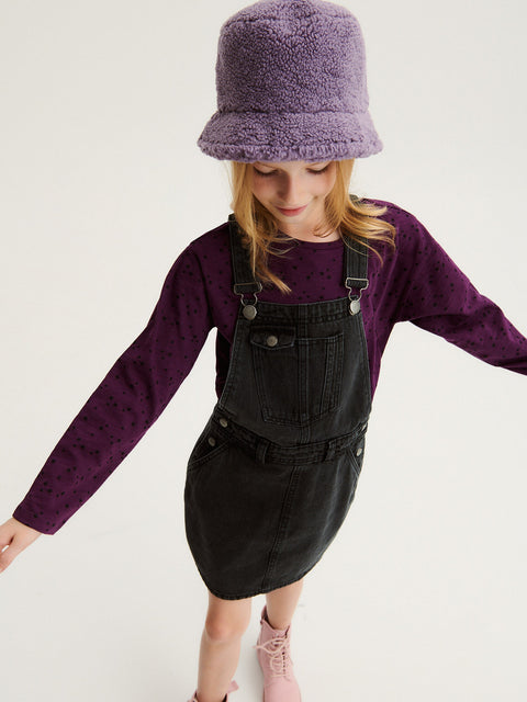 Person wearing a purple sweater, black overalls, and a purple bucket hat on a white background