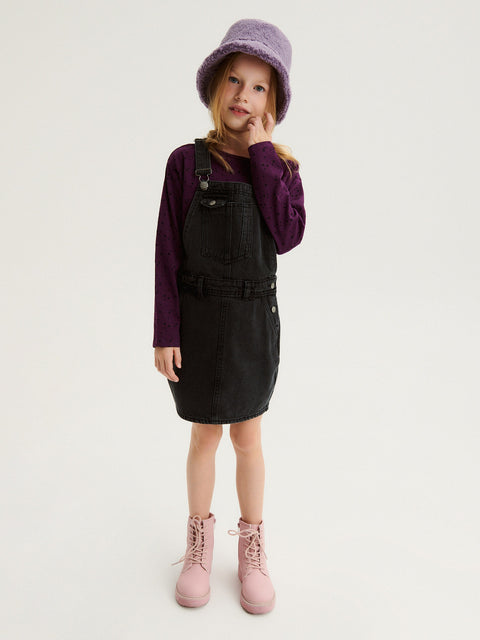 Young girl wearing a black pinafore dress, purple long-sleeve shirt, pink boots, and a purple hat on a white background