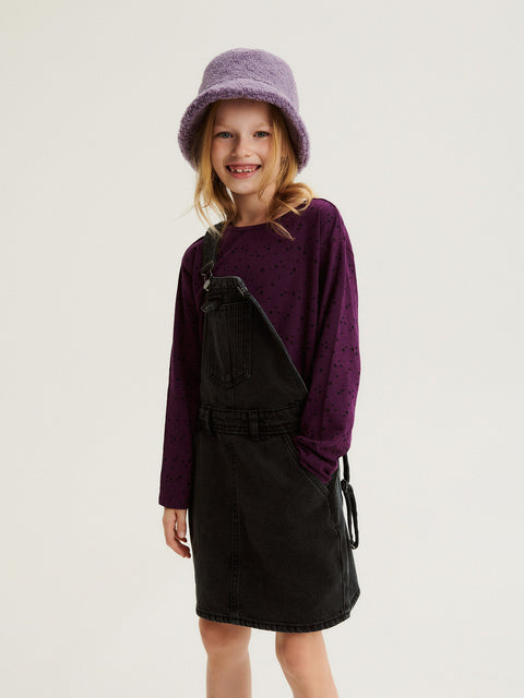 Young girl wearing a purple sweater and black pinafore dress with a matching hat on a white background