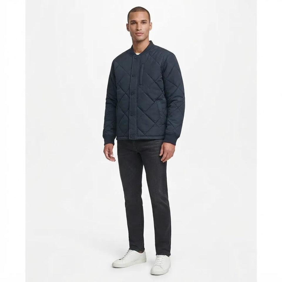 A man modeling a dark navy blue quilted jacket, paired with dark gray jeans and white sneakers, standing against a plain white studio background.