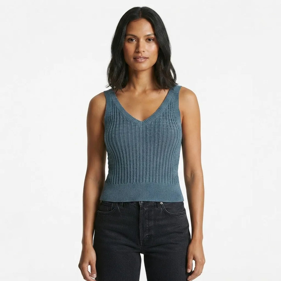 A woman modeling a sleeveless ribbed knit V-neck top in a muted blue-gray color, paired with dark jeans, against a plain white studio background.