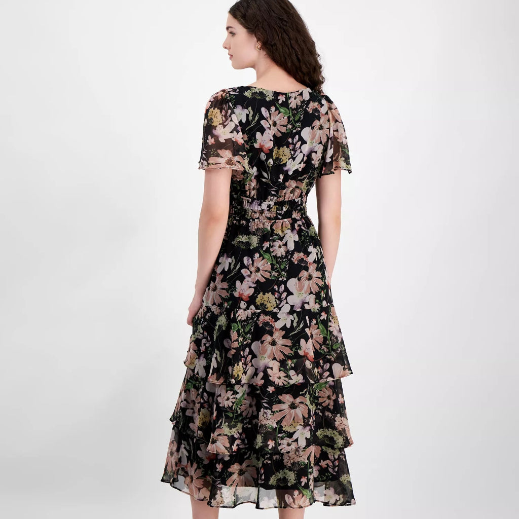A woman modeling a black midi dress with a pink and green floral pattern, shown from the back against a light gray studio background.