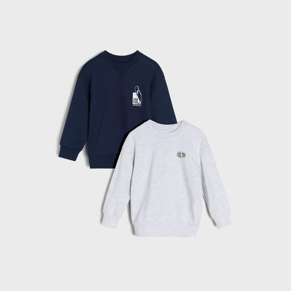 Two crewneck sweatshirts from the brand Chaps, displayed as a flat lay on a light gray background. The top sweatshirt is navy blue with a small white graphic on the left chest, and the bottom sweatshirt is light heather gray with a small metallic logo on the left chest.
