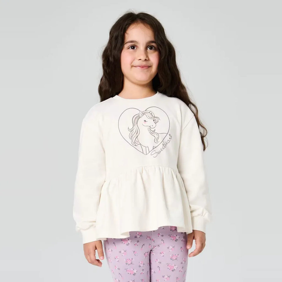 A young girl with long dark hair stands against a light gray studio background wearing a cream-colored long-sleeve top with a black line art graphic of a horse and the word 'Sofia' on the front, paired with light purple floral leggings.