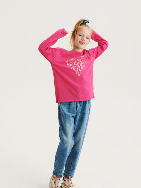 Child wearing a pink long-sleeve shirt with a heart design and blue jeans on a white background