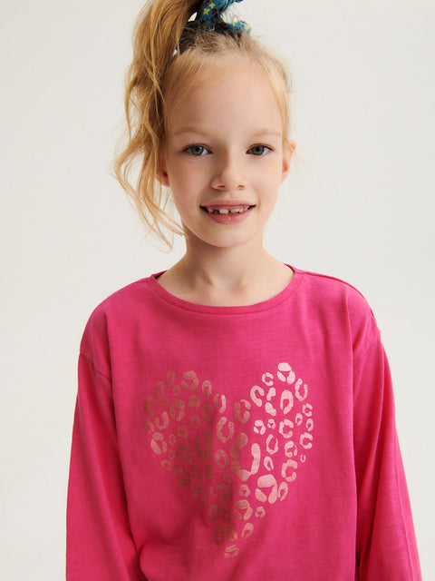 Young girl wearing a pink long-sleeve shirt with a heart design on a plain background