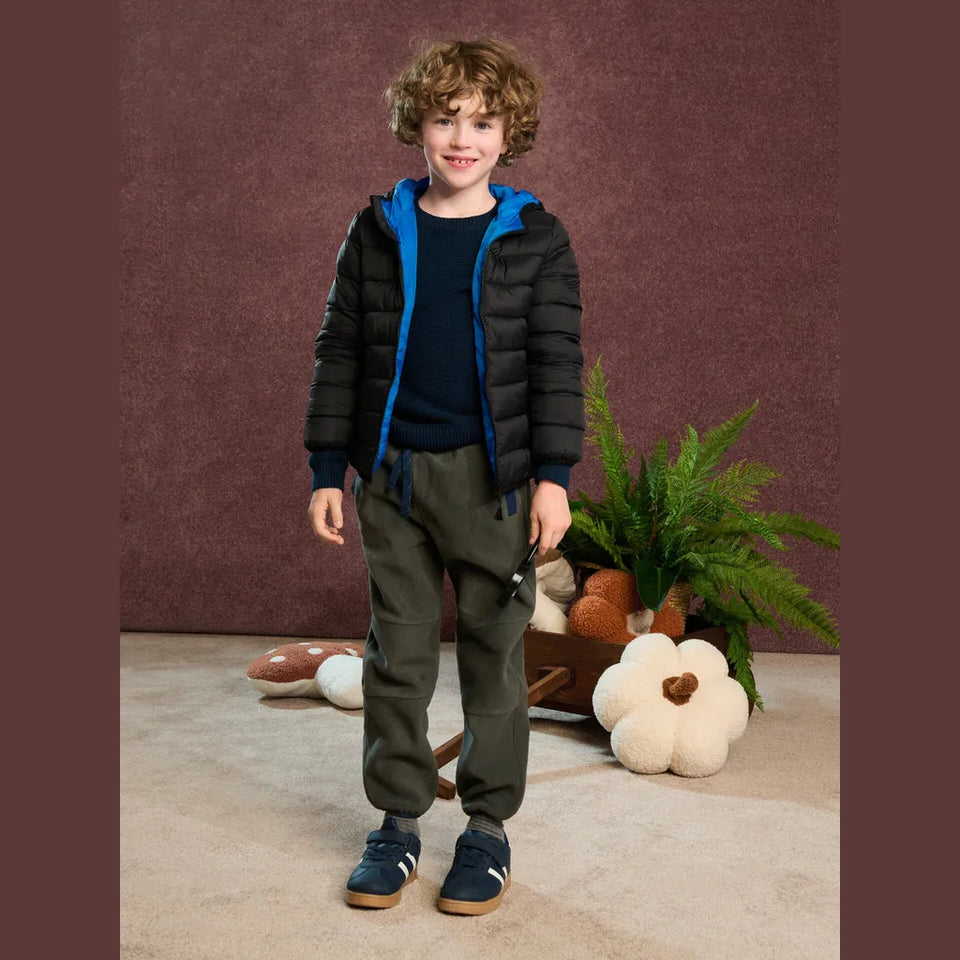 A young boy stands in a studio setting wearing a black puffer jacket with a bright blue hood and lining, a dark navy shirt, olive green pants, and navy sneakers. The background features a textured dark brown wall, a beige floor, and decorative props including a potted fern and pumpkins.