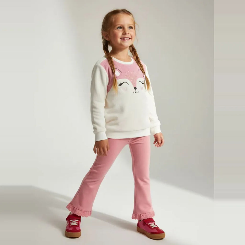 A young girl with braided hair stands in a studio wearing a white long-sleeve sweatshirt with a pink fox face graphic, pink pants, and pink shoes.