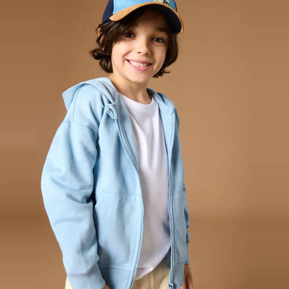 A smiling young boy wearing a light blue zip-up hoodie over a white t-shirt, paired with a blue and tan baseball cap, set against a solid warm brown studio background.