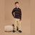 A young boy stands in a studio wearing a long-sleeve polo shirt with horizontal navy and brown stripes, beige corduroy pants, and navy sneakers. A beige pumpkin-shaped cushion sits on the floor beside him against a solid beige backdrop.