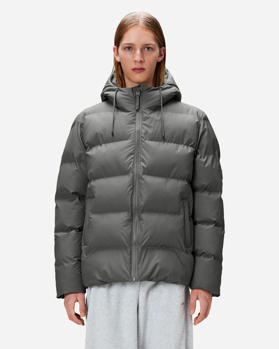 A model wearing a dark gray hooded puffer jacket with horizontal quilting and a front zipper, paired with light gray sweatpants, against a plain light gray background.