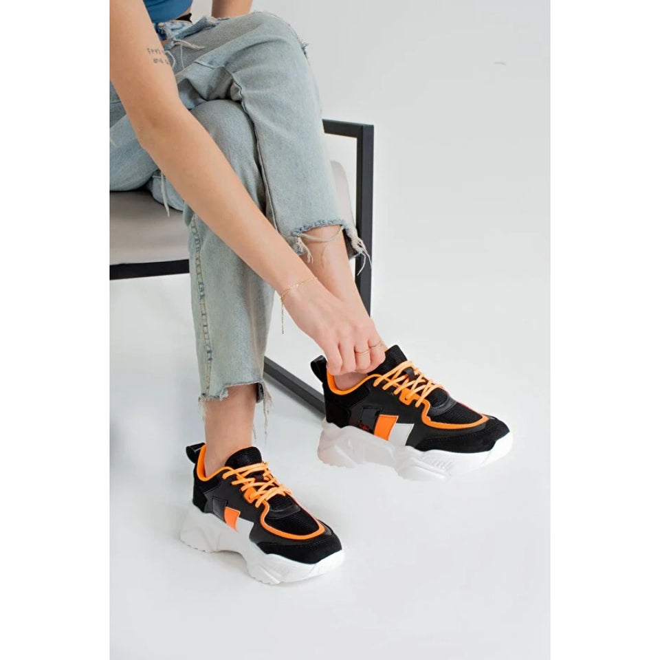 A person seated on a chair wearing black chunky platform sneakers with bright orange laces and orange side accents, paired with light blue frayed-hem jeans in a clean studio setting.