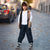 A young boy stands on a cobblestone street wearing a beige hooded puffer vest over a white sweatshirt, dark cargo pants, and black-and-white sneakers.