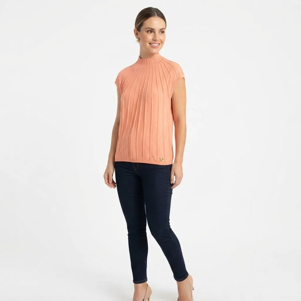 A woman modeling a sleeveless, high-neck ribbed knit top in a muted peach-pink color, paired with dark navy skinny jeans and beige heels, against a plain white studio background.