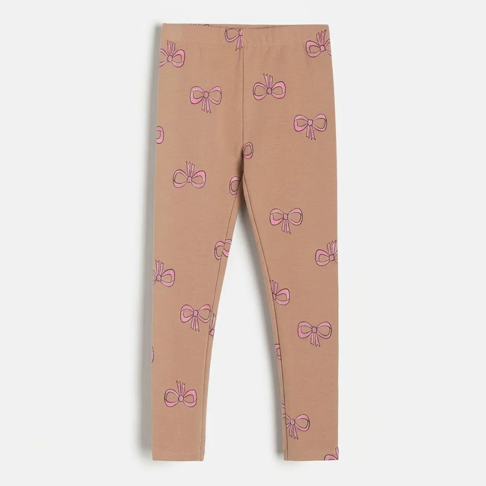 A pair of children's leggings in a dusty rose-beige color, featuring an all-over pattern of small pink bows outlined in darker pink, shown flat against a light gray background.