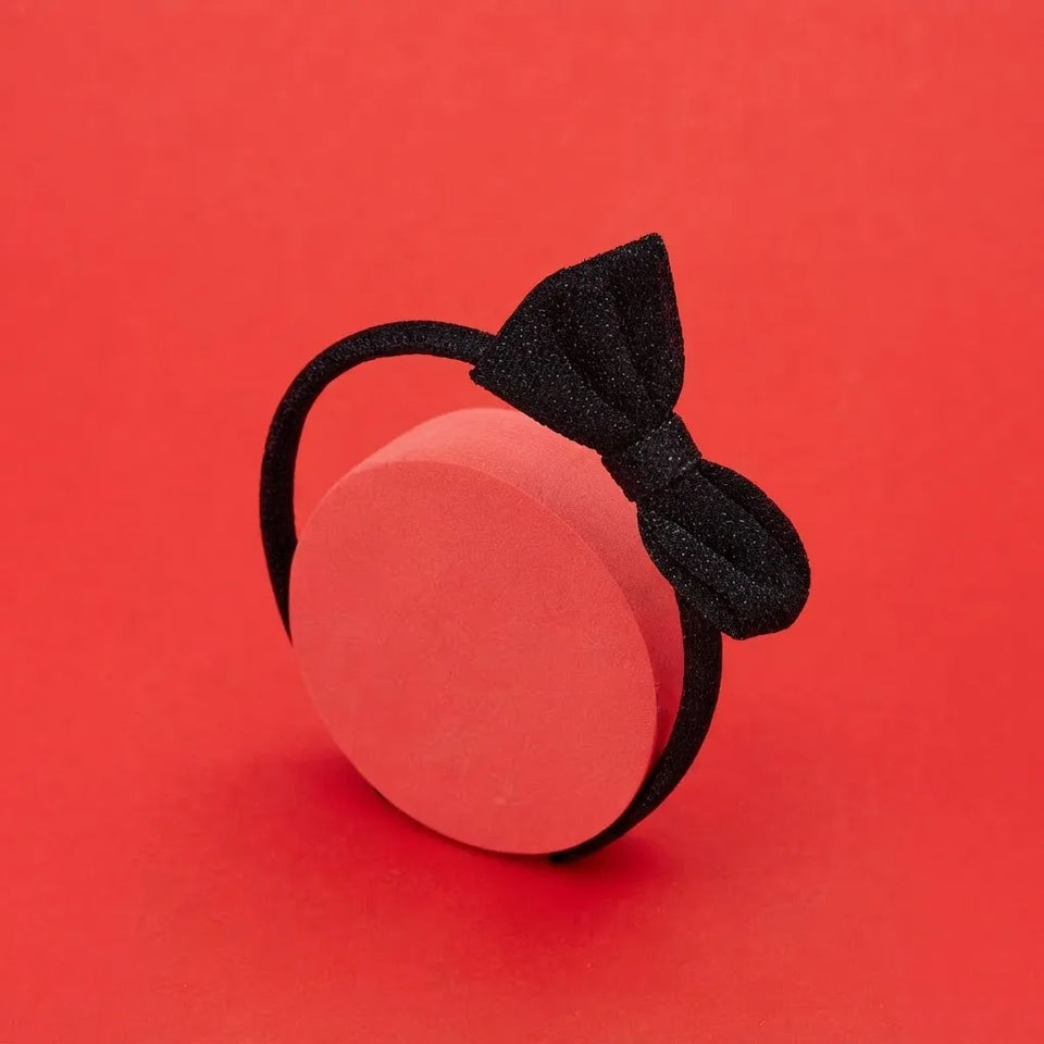 A black elastic hair band with a glittery black bow, displayed on a round pink display block against a solid red background.