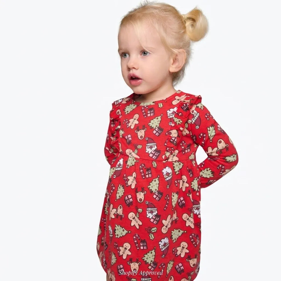 A toddler girl with blonde hair in a ponytail wearing a red long-sleeve dress with a gingerbread man and Christmas tree pattern, photographed against a light gray background.