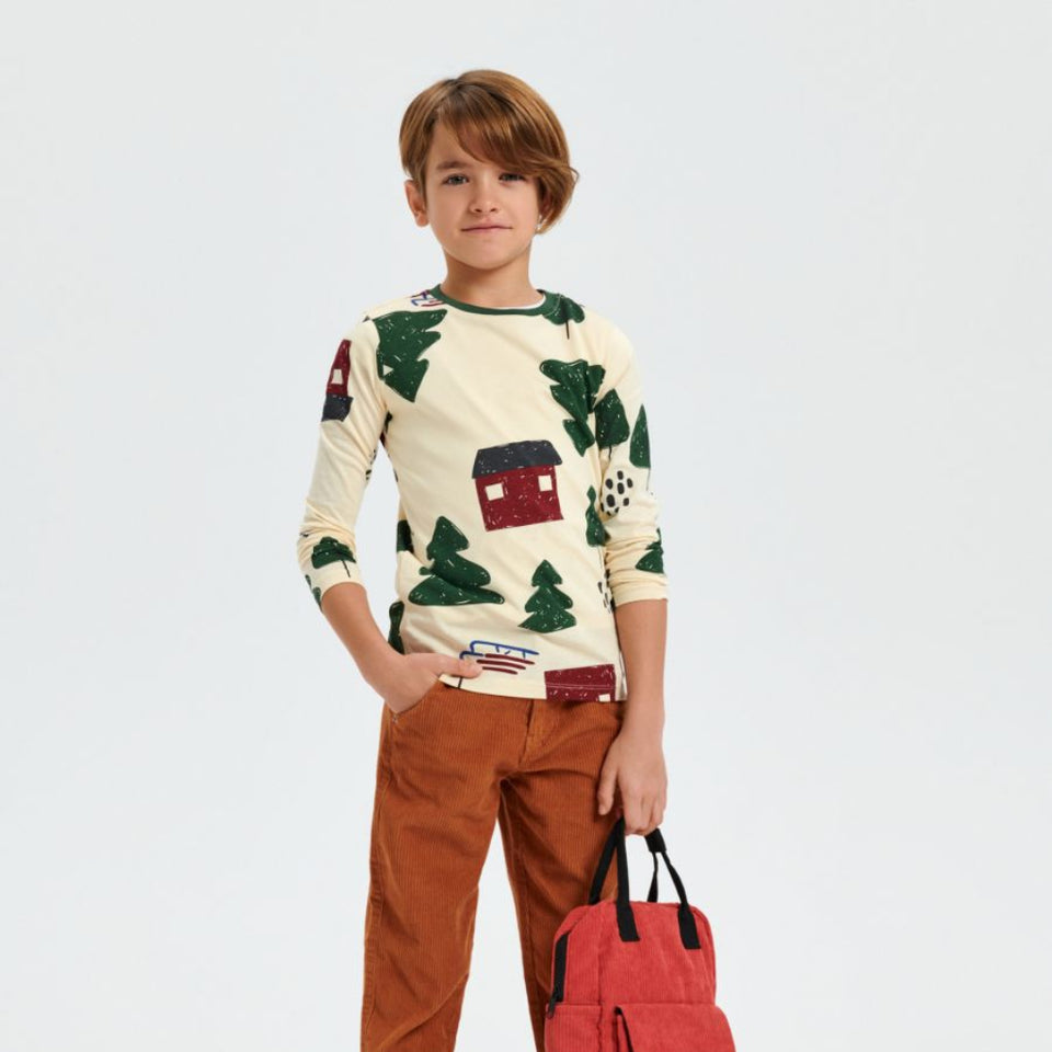 A young boy modeling a cream long-sleeve shirt with a green pine tree and red house pattern, paired with rust-brown pants, black sneakers, and holding a red backpack against a light gray studio background.