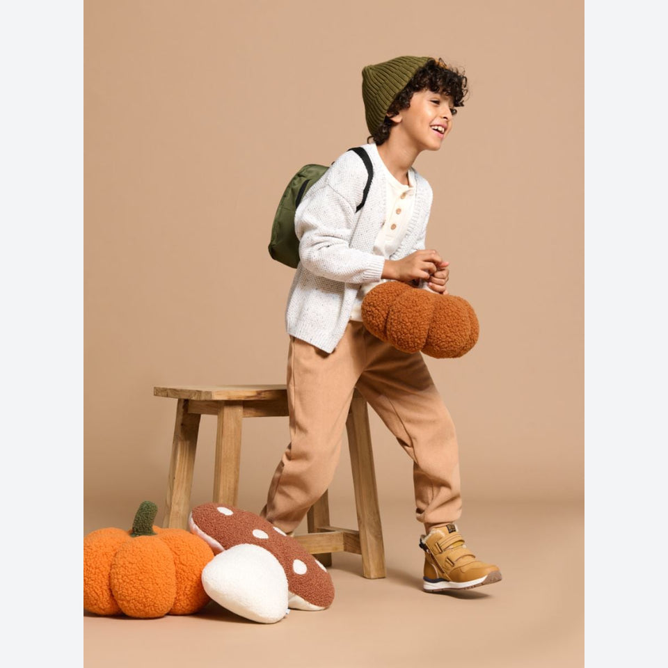A child wearing a green knit beanie, white cardigan, tan pants, and tan boots stands on a wooden stool in a studio setting, holding a brown fuzzy pumpkin-shaped pillow. More matching pillows are on the floor, and the background is a solid warm beige.