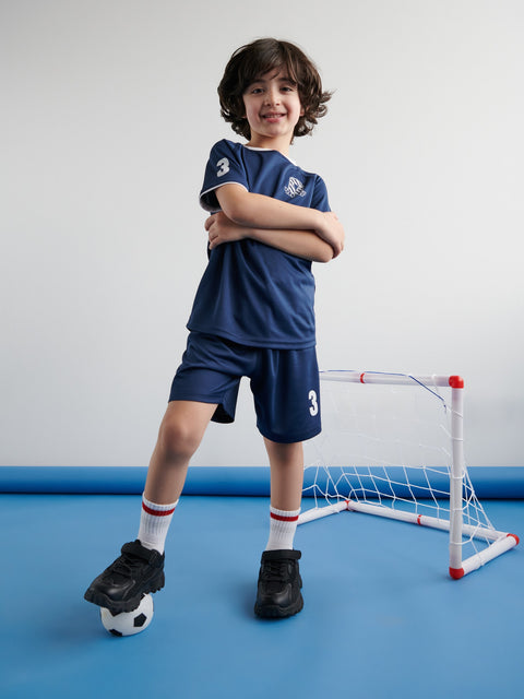 Child wearing a blue sports outfit with number 3, standing on a blue floor with a small soccer goal in the background.
