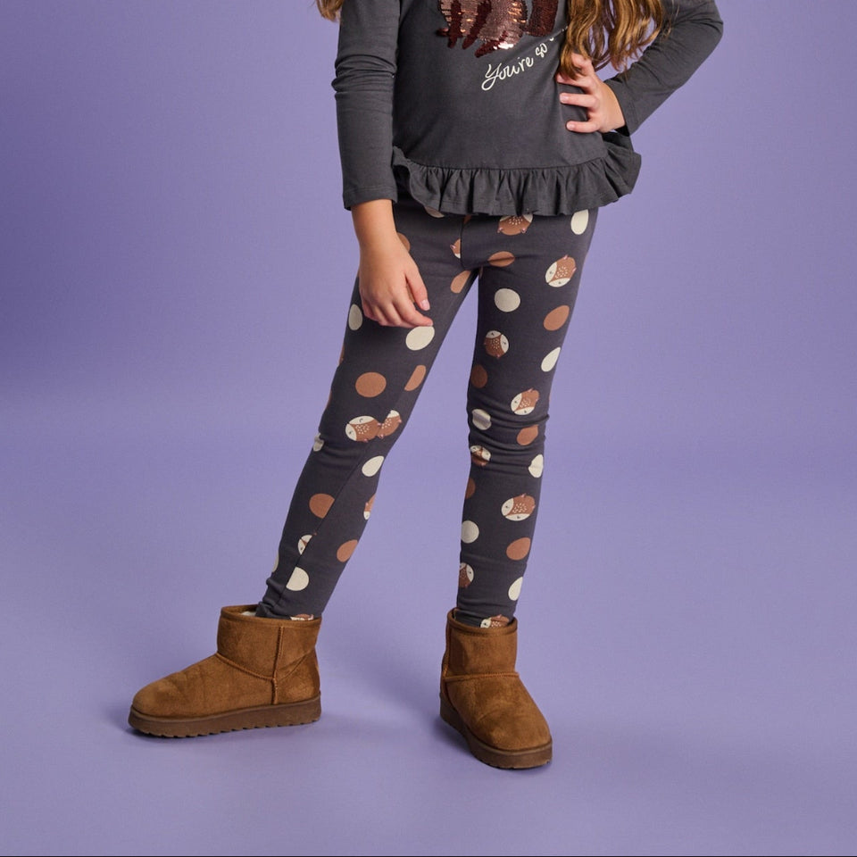 A child model wearing dark gray leggings with a brown and cream polka dot pattern, styled with a dark gray ruffled long-sleeve top and brown suede boots against a solid lavender studio background.