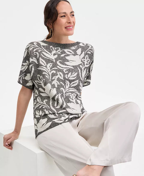Woman wearing a patterned top and white pants sitting on a white surface.