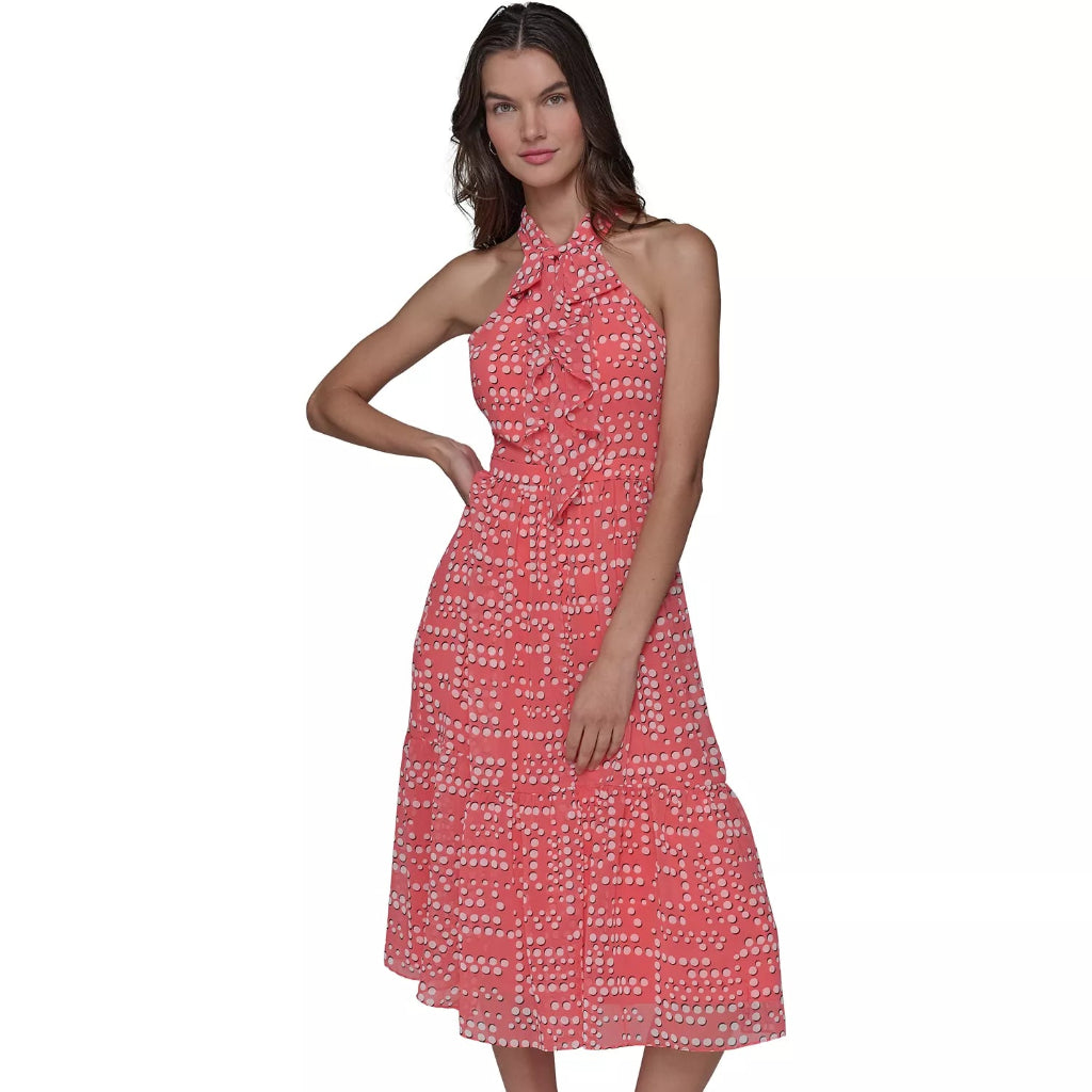 A woman modeling a coral pink halter midi dress with a white abstract pattern, paired with black strappy high-heeled sandals, against a plain white background.