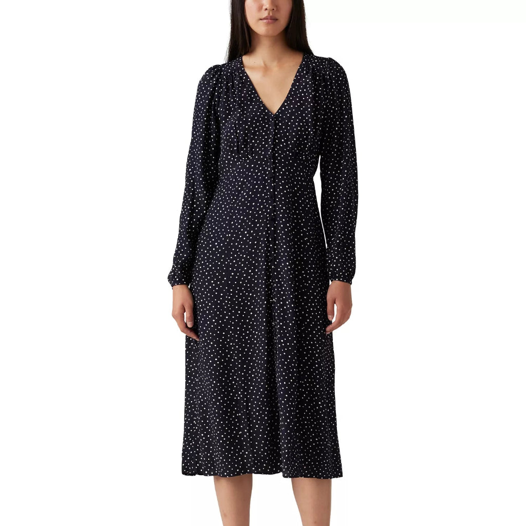 A woman modeling a long-sleeve midi dress with a dark navy base and a dense white micro-dot pattern, paired with black strappy sandals against a plain white background.