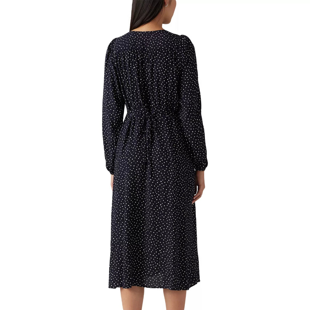 A woman viewed from the back wearing a dark navy midi dress with a small white polka dot pattern, long sleeves, and a tie at the back waist, paired with black pointed-toe heels against a plain white background.
