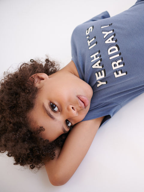 Child wearing a blue t-shirt with 'YEAH IT'S FRIDAY!' text, lying on a white surface.