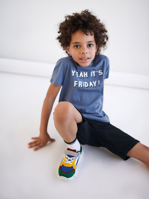 Child wearing a blue t-shirt with 'Yeah It's Friday!' text, sitting on a white floor.