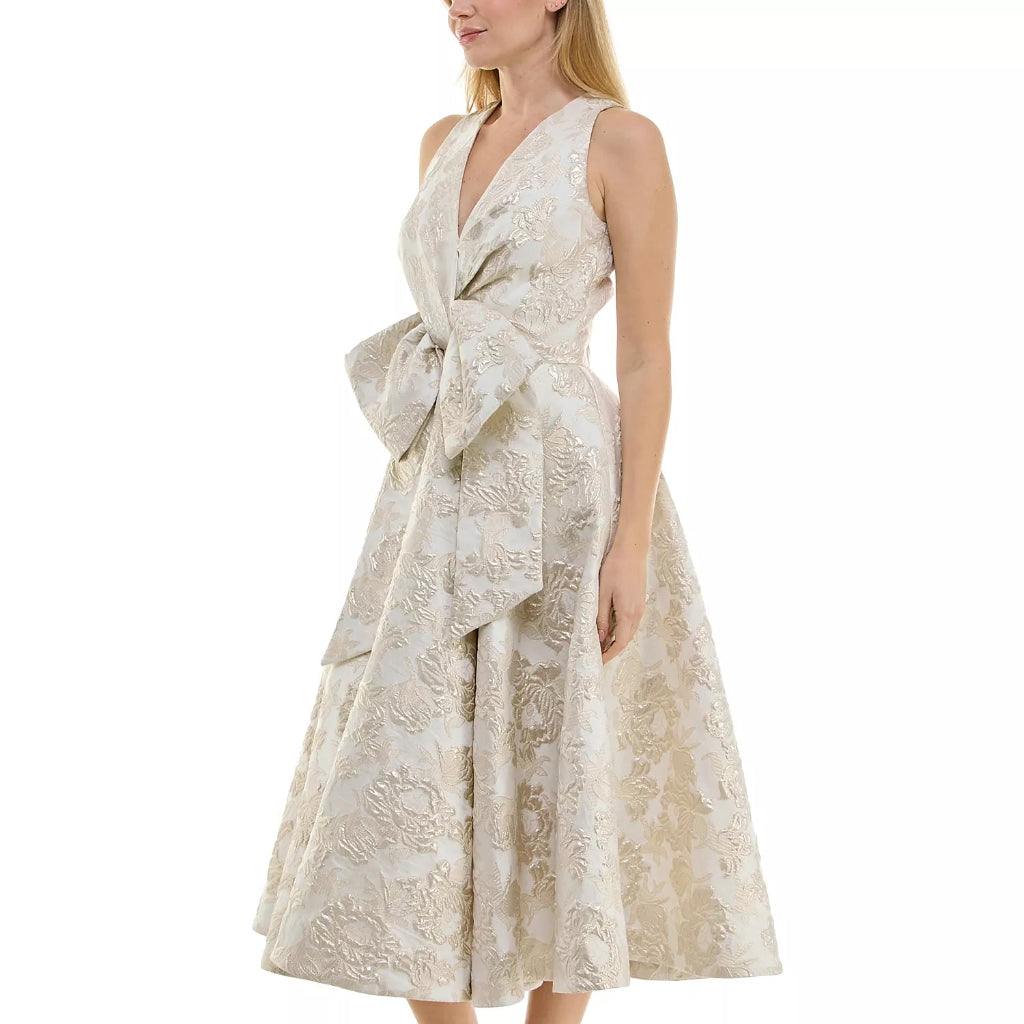A woman models a sleeveless midi dress with a deep V-neckline, a large twisted waist bow, and a tiered skirt. The dress features a textured beige and cream floral jacquard pattern.