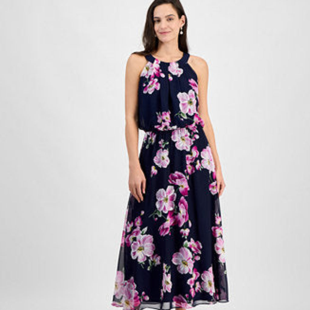A woman modeling a sleeveless navy maxi dress with a pink and white floral print, paired with white heeled sandals, against a light gray studio background.