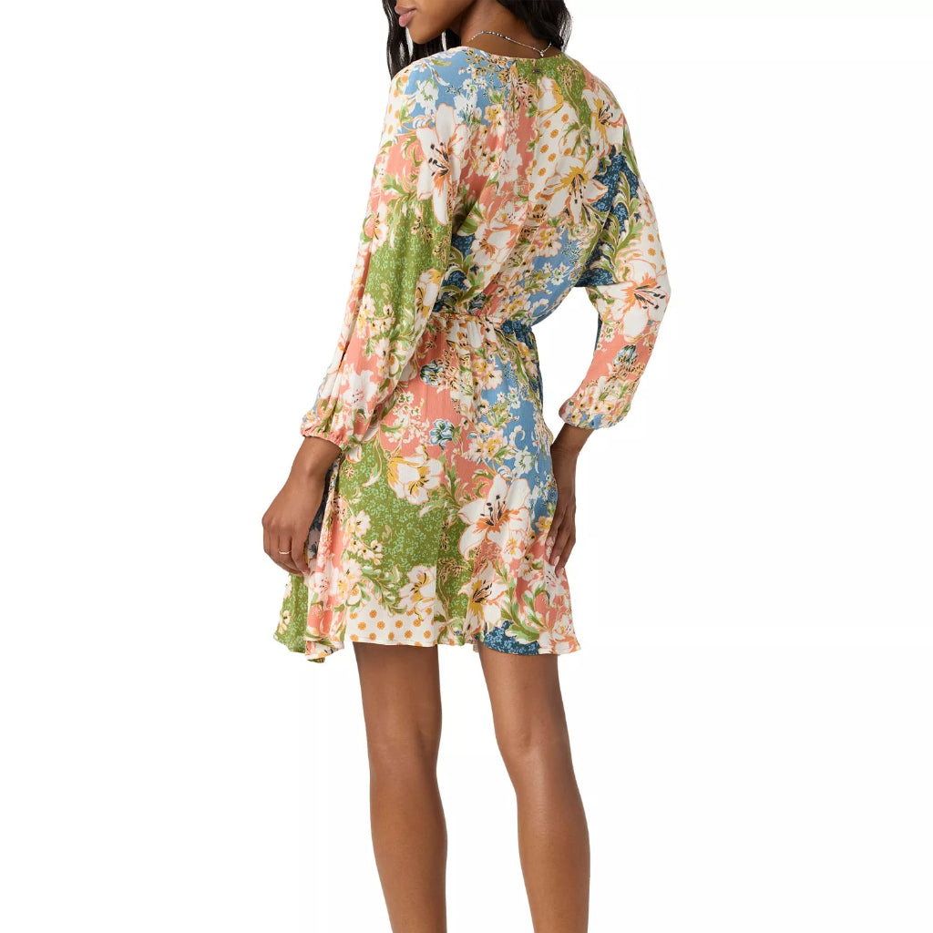 A woman modeling a long-sleeve mini dress with a colorful floral print, shown from a side-back angle against a plain white background.
