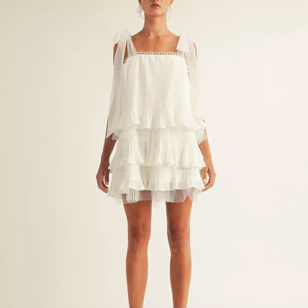 A model wearing a white tiered ruffle mini dress with cold-shoulder sleeves and light blue strappy heels, shot against a plain off-white studio background.