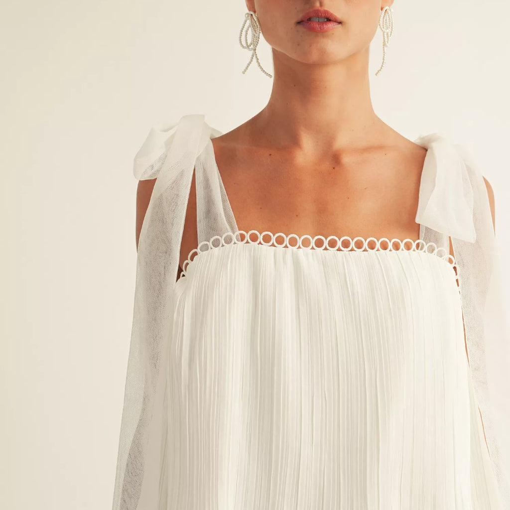A close-up of a model wearing a white cold-shoulder top with sheer flutter sleeves, a square neckline accented by a ring trim, and a pleated bodice.
