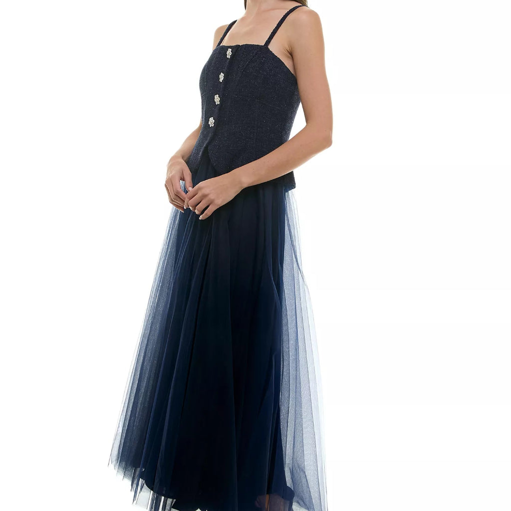 A woman modeling a sleeveless navy maxi dress with thin straps, a textured bodice with decorative buttons, and a layered tulle skirt, shown against a plain white background.